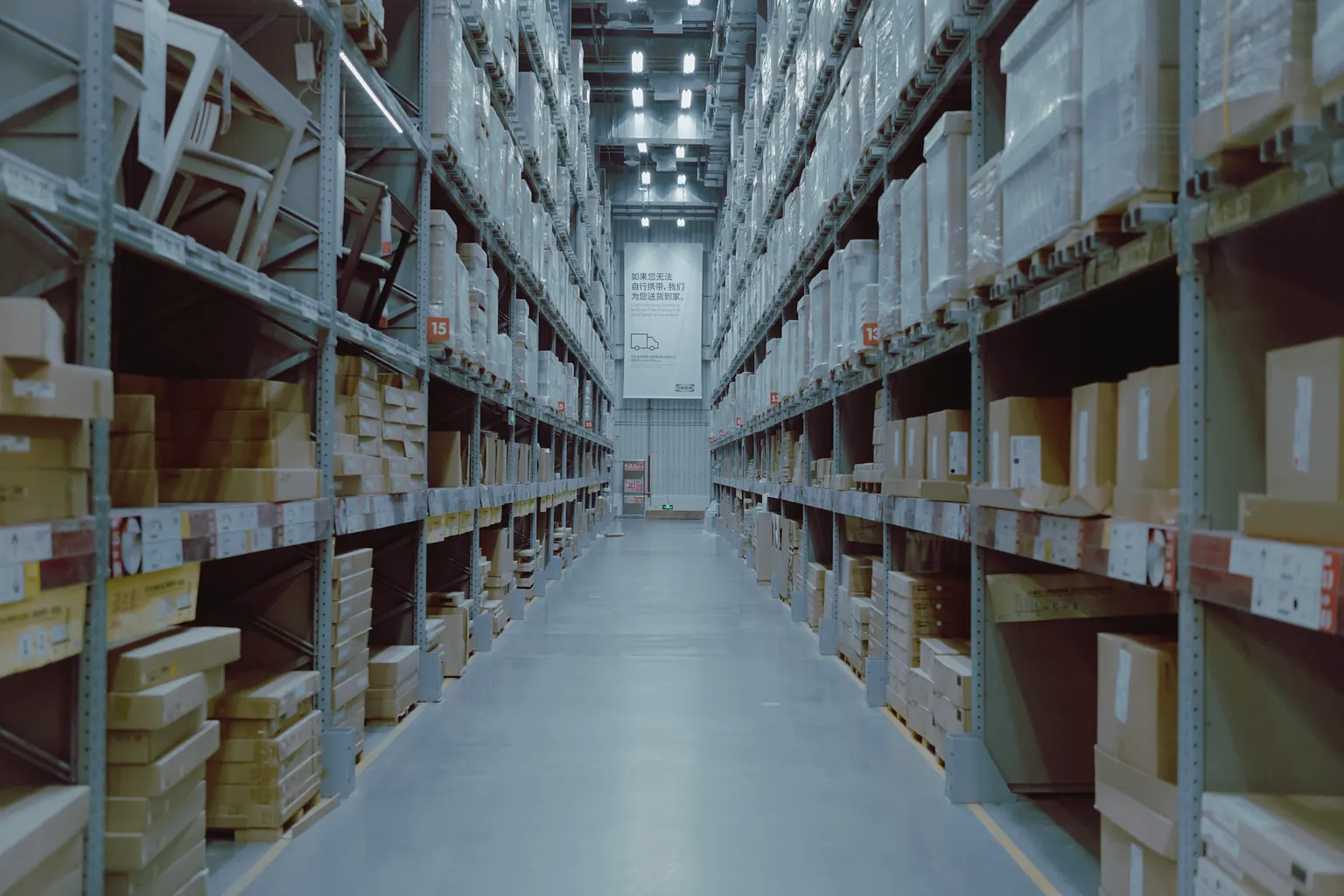 A long warehouse aisle between tall shelves of boxed stock under bright lights.
