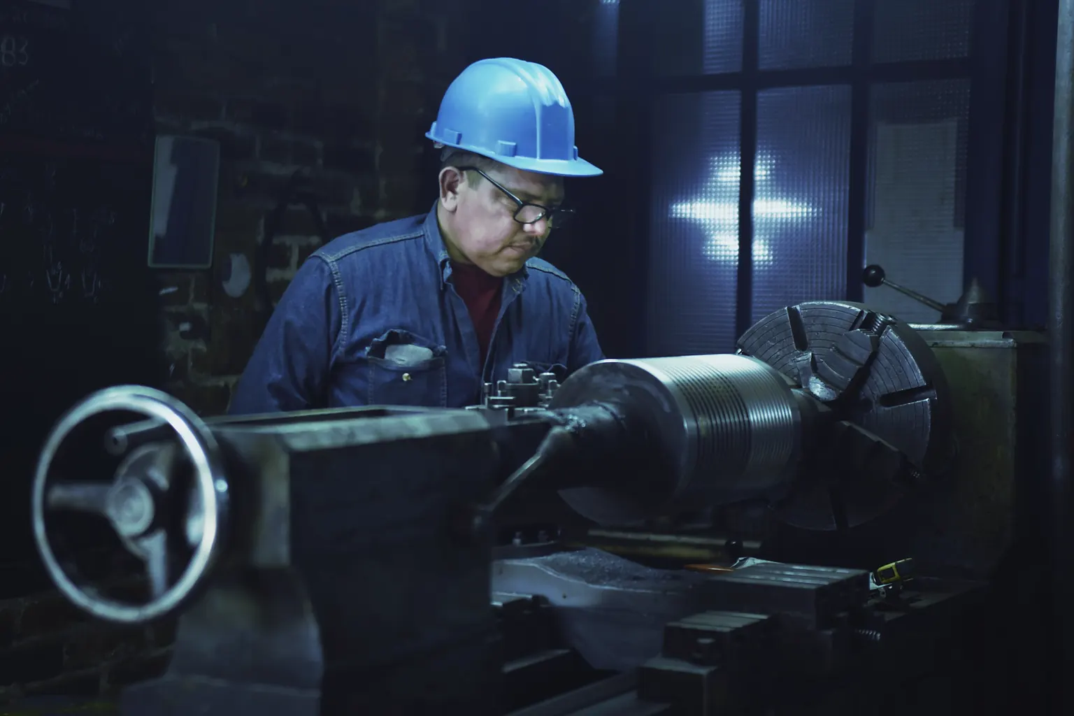 A machinist in a blue hard hat and overalls works at a lathe in a dim workshop.